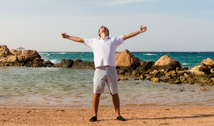homem jpvem com os braços abertos feliz na beira de uma praia com rochas atrás