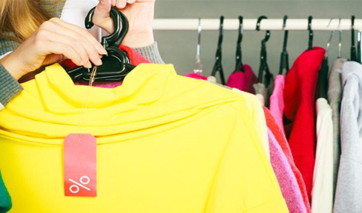 Na foto há uma mulher segurando cabides com um roupa amarela com uma etiqueta de promoção vermelha. Atrás dela há mais roupas.
