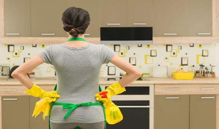 Na foto há uma mulher com luvas de borracha amarelas e avental verde olhando para a pia de uma cozinha. Ela está de costas.