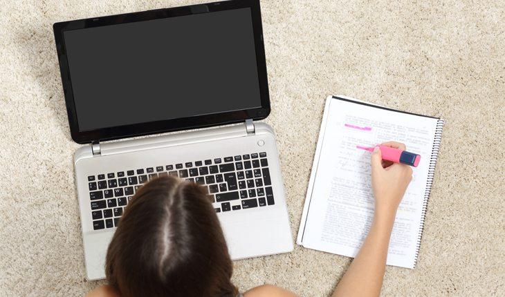 Na foto há uma menina estudante com um computador e um caderno. A foto é vista de cima.