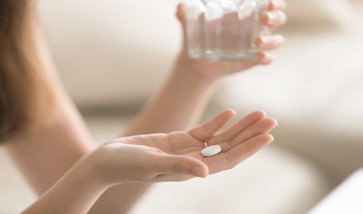 Na foto há a mão de uma pessoa com uma pílula branca de remédio e a outra mão está segurando um copo de água.