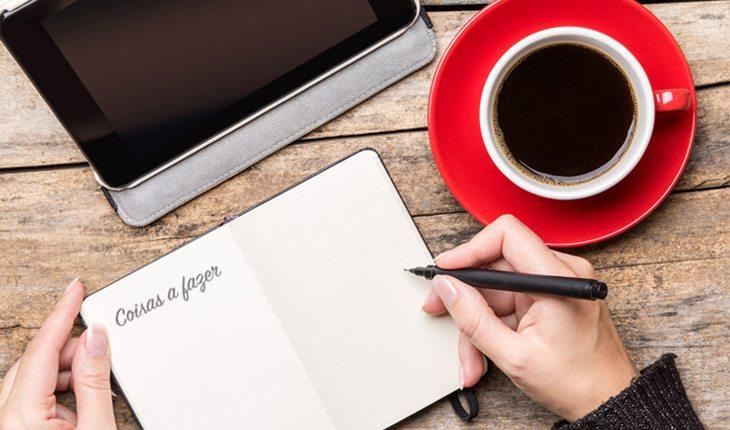 Na foto há uma pessoa com uma caneta na mão em cima de um livro branco e próximo a um computador e uma xícara de café.