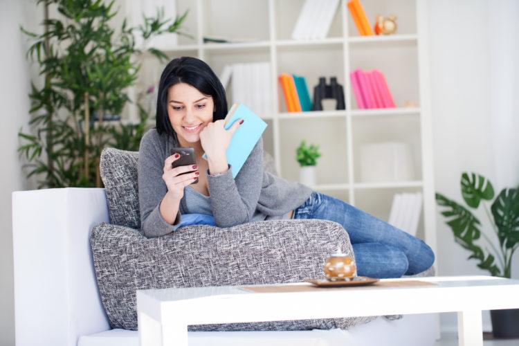 mulher, sentada, mechendo no celular, livro, sozinha, sofá, sorrindo, iphone e ipad