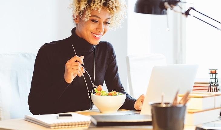 A foto mostra uma mulher tomando café da manhã enquanto mexe no computador quebrando a rotina