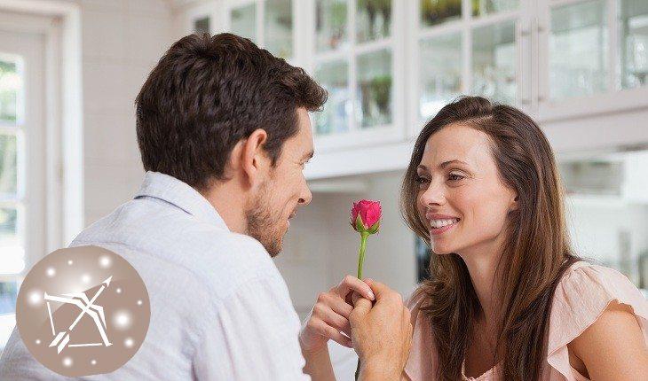 Na foto há um casal heterossexual e o homem está dando uma rosa para a mulher.
