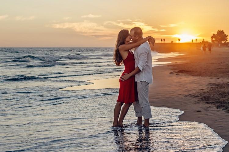 Na imagem, o casal está na beira do mar se beijando, ela com um vestido vermelho e ele com roupa branca no por do sol. Amor e atração. 