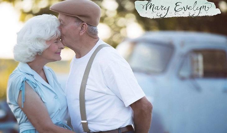 A foto mostra um casal de idosos. O homem está beijando a testa da mulher