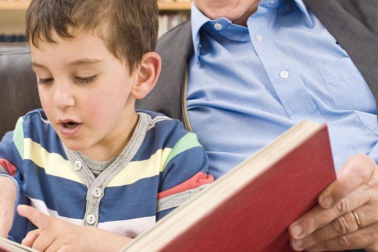 A foto mostra um menino lendo um livro junto com seu avô