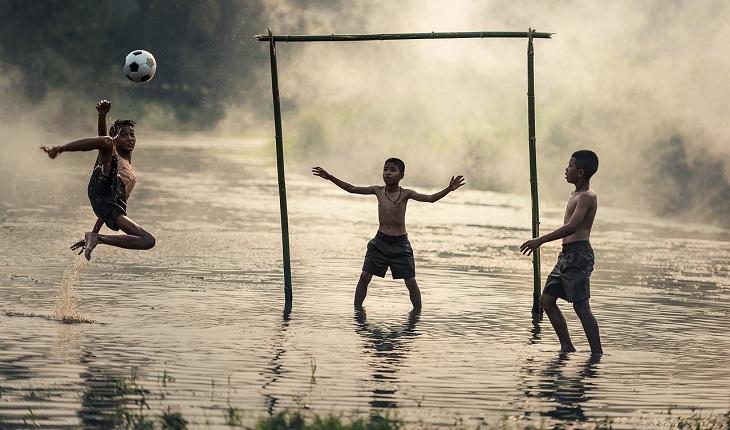 Na imagem, três meninos carentes jogam futebol em um campo alagado com o gol improvisado de madeira. Rainha da Paz.