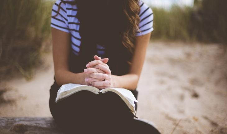 Na imagem, uma mulher está sentada com a biblia aberta e as mãos fechadas em oração. Rainha da Paz.