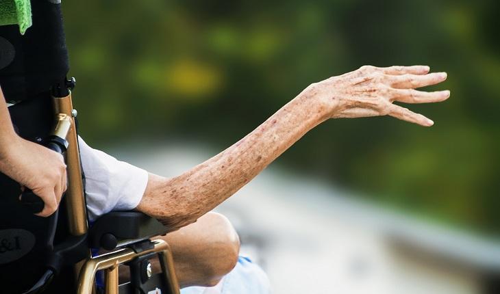 Na imagem, o braço de uma mulher enferma e idosa está estendido na cadeira de rodas que está sendo empurrado por alguém. Rainha da Paz.
