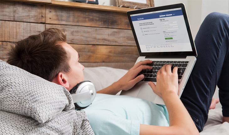 homem deitado na cama, segurando um notebook conectado ao Facebook