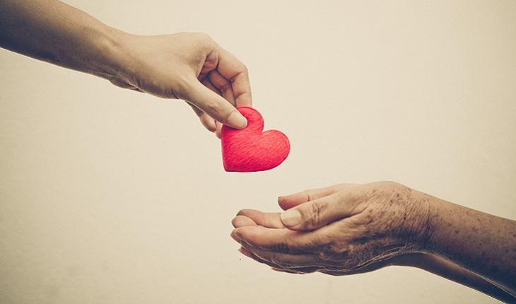 A foto mostra uma mão de uma pessoa jovem entregando um coração de papel para uma mão de uma pessoa idosa. A imagem faz referência aos cuidados necessários com o coração para quem tem obesidade na terceira idade