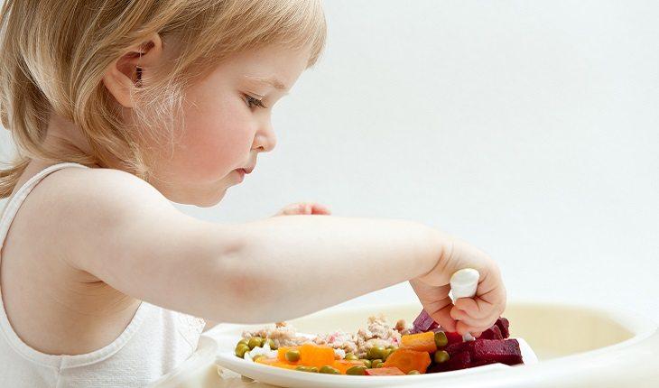 A foto mostra uma menina loira comendo frutas com a mão