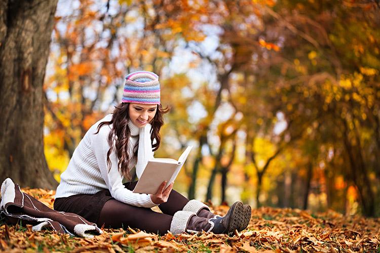 mulher com roupa de frio lendo sentada em folhas secas em um campo com árvores