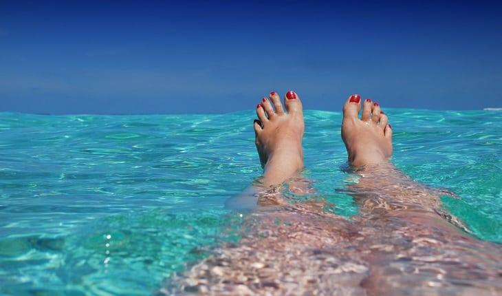 Na imagem, uma mulher está com as pernas mergulhadas no mar os pés para fora. Área íntima feminina.
