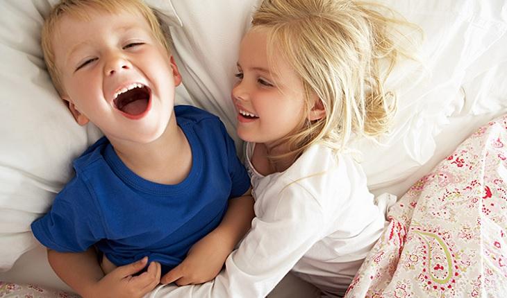 A foto mostra duas crianças deitadas em uma cama gargalhando. Dar risada funciona como um analgésico natural