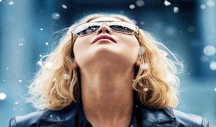 A atriz Jeniffer Lawrence olha para cima, com os cabelos curtos e óculos de sol, flocos de neve desfocados caem do céu