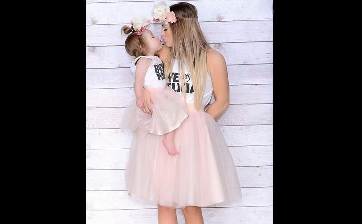Mulher e filha bebê, dão um beijo. As duas estão vestidas com camisetas brancas estampadas e uma saia plissada de balé