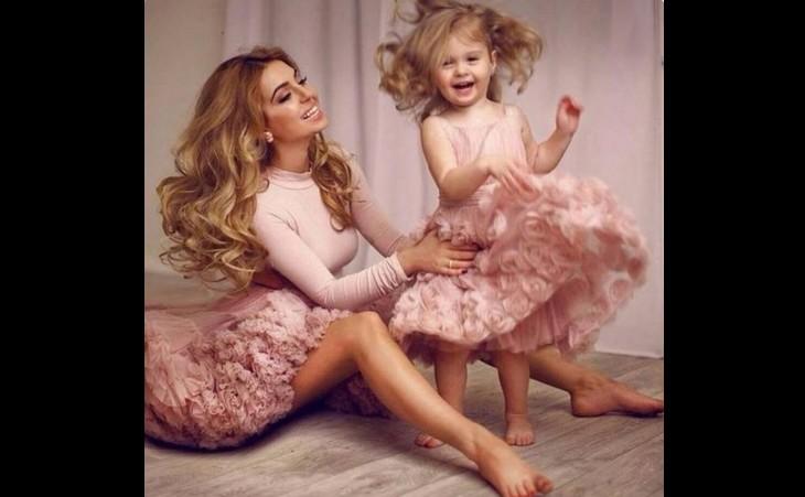 Mãe está sentada enquanto a filha criança está em pé dançando, as duas usam um vestido rosa