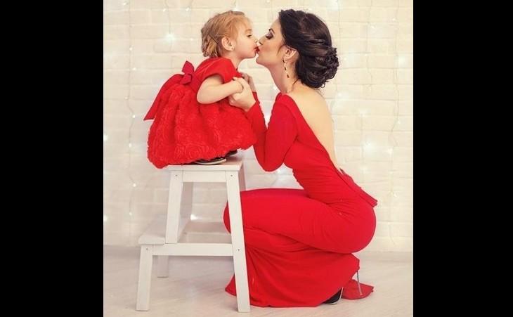 Mãe está agachada dando um beijo na filha, que está em pé em um banquinho para ficar da altura dela, as duas usam vestidos vermelhos