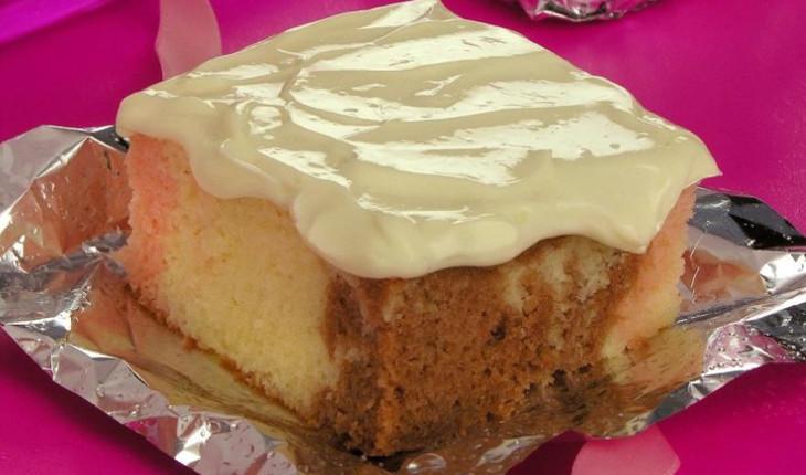 Na foto há um pedaço de bolo quadrado nas cores creme, marrom e rosa, sendo um bolo napolitano. Há um creme claro como cobertura e o pedaço está sobre um papel-alumínio.