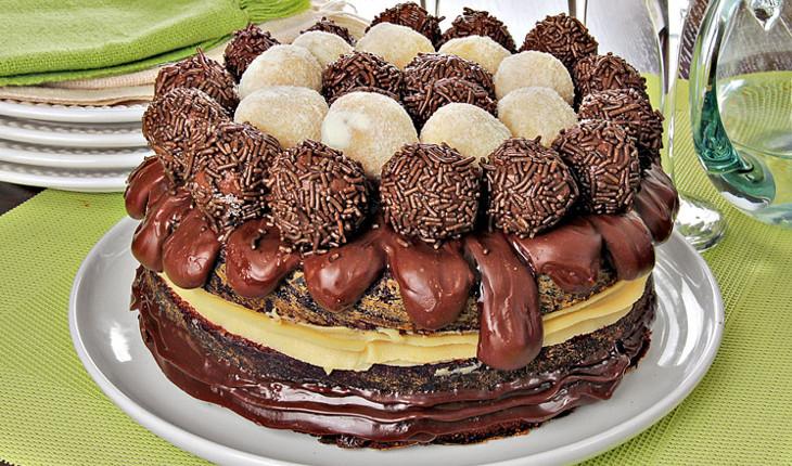 Na foto há um bolo inteiro de chocolate que é feito em várias camadas. No topo há diversos tipos de brigadeiro.