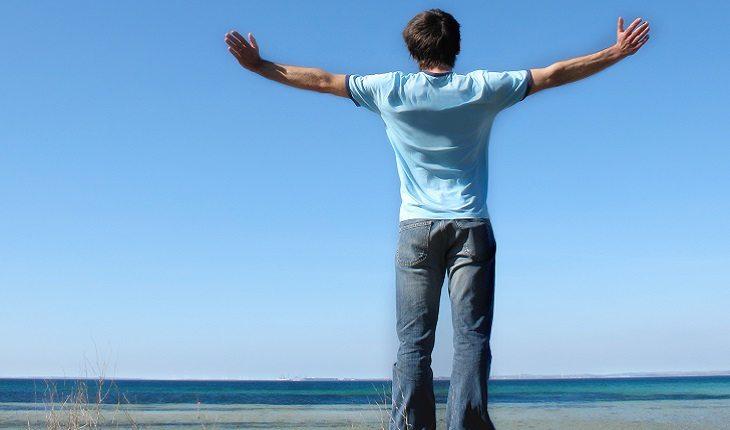 A foto mostra um homem com camiseta azul de braços abertos em frente ao mar
