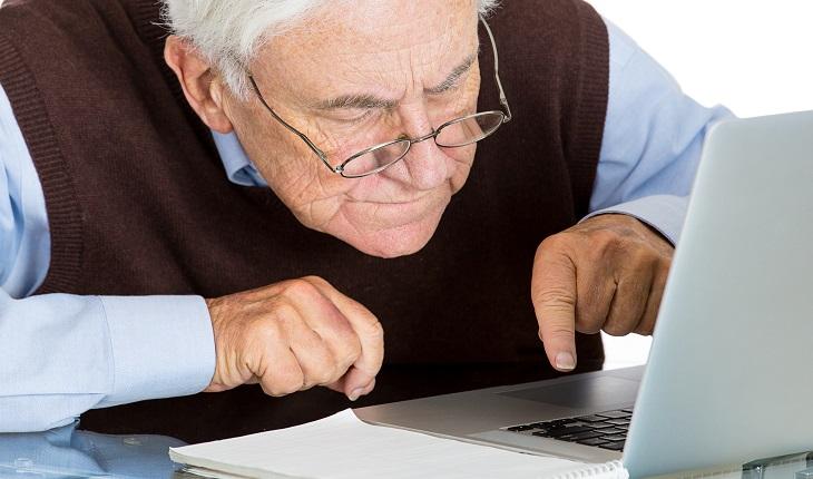 Na foto há um homem idoso mexendo no notebook com uma postura incorreta. Fazer a correção postural é importante para evitar dores nas costas na terceira idade