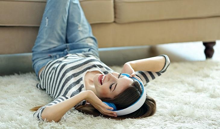 Na foto há uma mulher deitada no chão com os pés no sofá ouvindo música com um fone. Ela está sorrindo