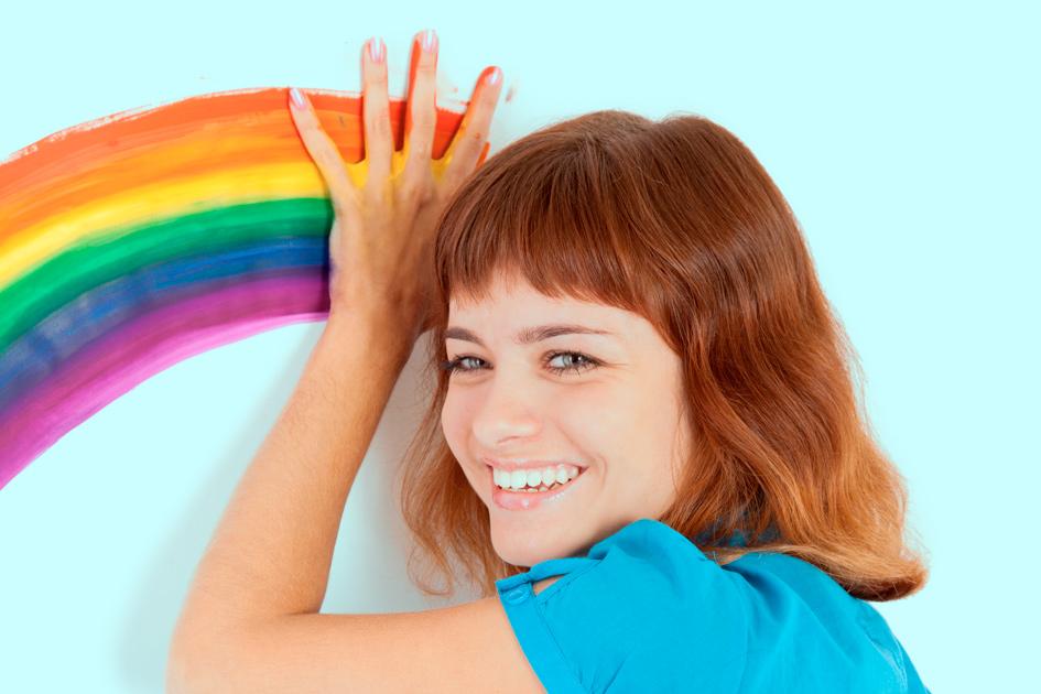 mulher ruiva de cabelo curto e blusa azul com a mão no arco-íris na parede