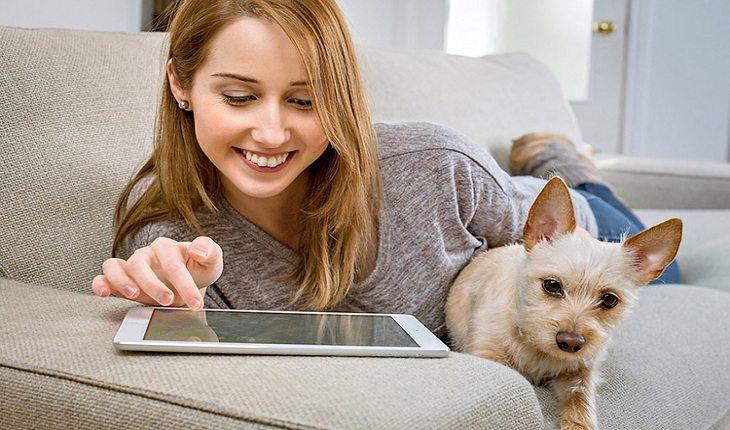 Mulher deitada em um sofá, mexendo em um tablet e ao lado de um cachorro