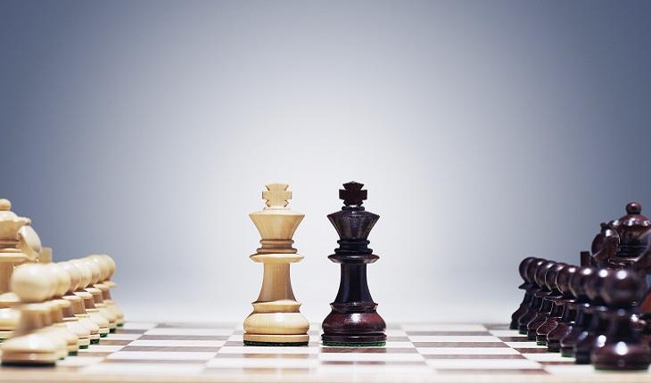 A foto mostra um tabuleiro de xadrez com duas peças uma de frente para a outra, uma branca e outra preta