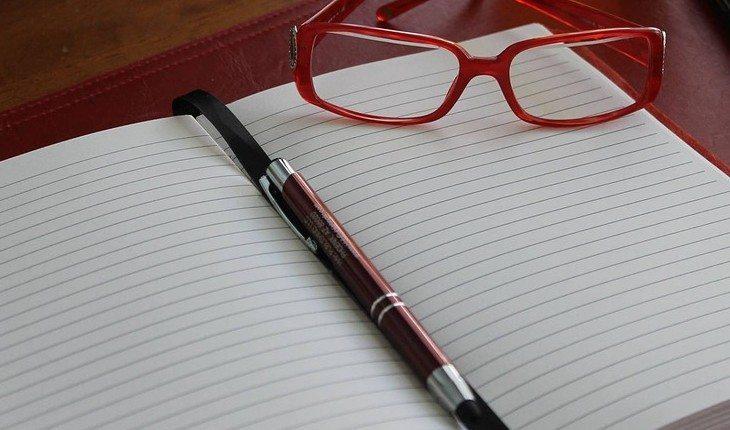 Na foto há um caderno em branco com uma caneta e um óculos vermelho em cima.