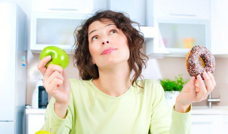 Na foto há uma mulher pensando se irá comer uma fruta ou uma rosquinha