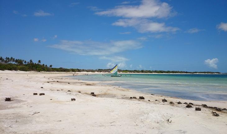 Vista da praia de Jericoacoara: uma área plana com areia clara de um lado e o mar com água azul clara do outro, com um barco de pesca centralizado.