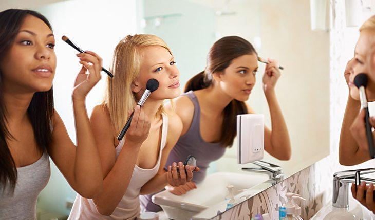 Na foto há 3 mulheres se maquiando olhando em um espelho