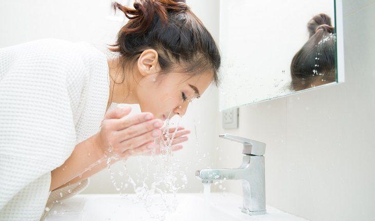 Na foto há uma mulher lavando o rosto em uma pia de banheiro