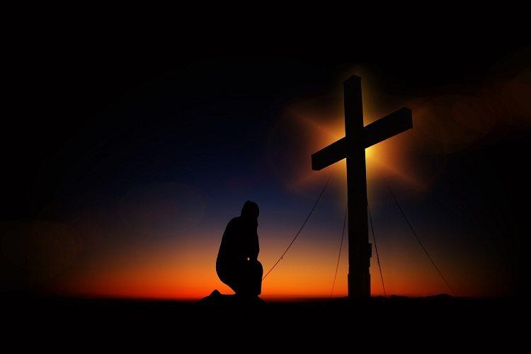 Figura humana ajoelhada rezando a prece do trabalhador em frente à cruz