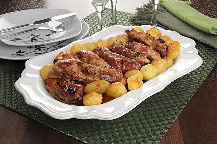 Costelinha ao forno com batata em uma travessa branca com pratos e taças ao fundo.
