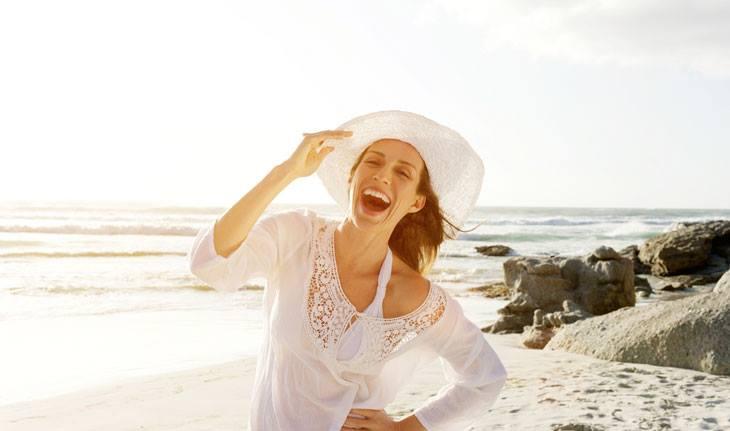 Mulher sorrindo, segurando chapeu em praia