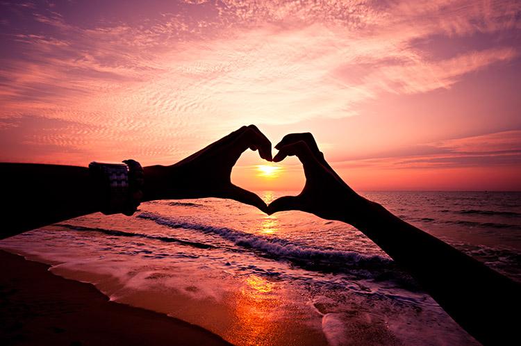 Casal fazendo coração com as mãos em frente ao por do sol em uma praia