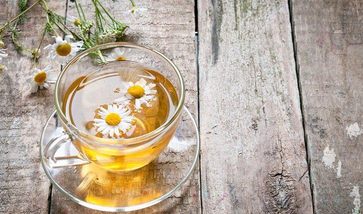 A foto mostra uma xícara com chá de camomila com a erva ao lado