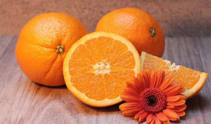 Na foto há algumas laranjas inteiras e um cortada ao meio e junto delas há uma flora laranja.