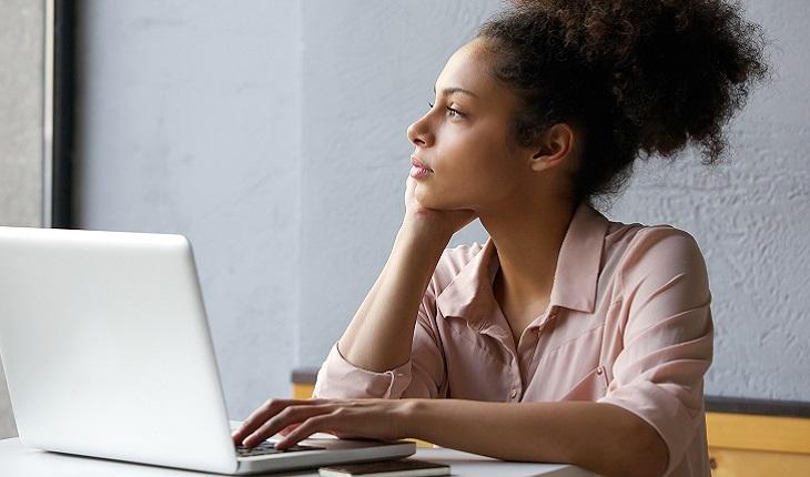 A foto mostra uma mulher negra sentada em frente a sua mesa de trabalho com um notebook. Ela está pensando em como criar novos hábitos
