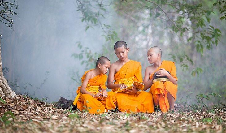 A foto mostra três meninos monges lendo um livro em um bosque. Escolas budistas