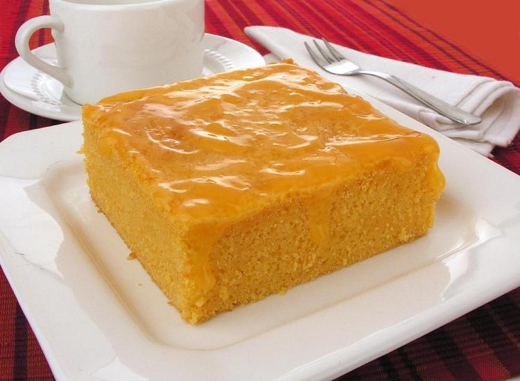 Na foto, um pedaço do bolo cremoso de tangerina está disposto em um prato de vidro quadrado. Em cima do bolo, está uma calda bastante cremosa. Na ambientação, a mesa está com uma toalha vermelha, e ao fundo estão uma xícara e um garfo.