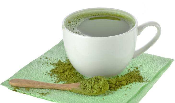 Xícara branca com chá verde e pó de chá verde em uma colher e ao redor, todos sobre um guardanapo verde
