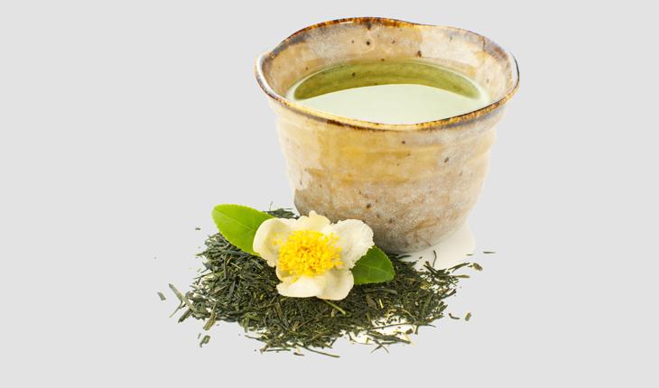 Chá verde japonês servido em em copo de cerâmica, ervas e uma flor branca decorando ao redo do copo e fundo cinza