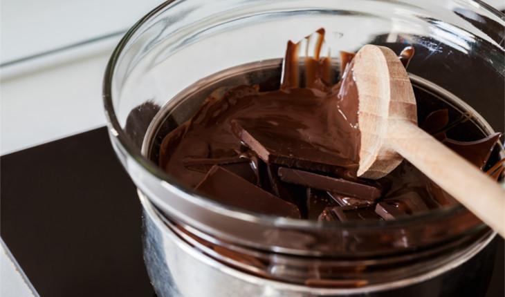 Na foto há um bowl de vidro cheio de chocolate que está sendo derretido em banho-maria com o auxílio de uma colher de pau.